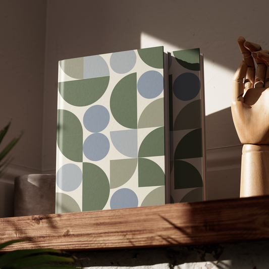 Two patterned books on a wooden surface with a hand and plant in the background
